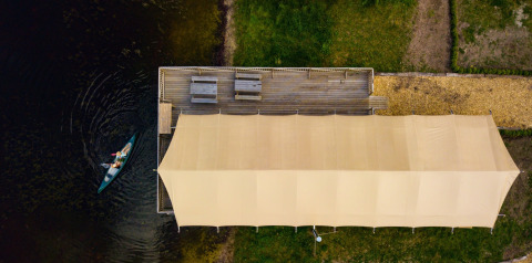 Vue aérienne d'une tente safari au bord d’un lac, une canoë et des bancs sur une terrasse en bois.
