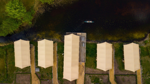 Vista aérea de tiendas safari junto a un río con senderos, vegetación y una canoa en el agua.