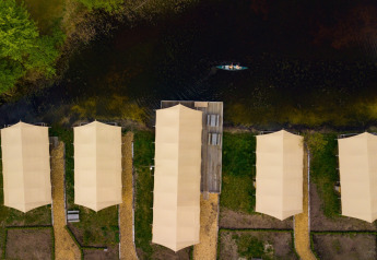 Vista aerea di tende safari allineate lungo un fiume con sentieri, vegetazione e una canoa sull’acqua.