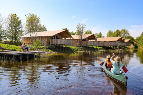 Two people paddle a canoe on a lake in front of safari tents, while others relax on a wooden dock.