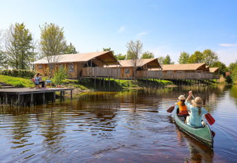 Due persone remano in canoa davanti a tende safari sul lago, altre si rilassano su un pontile di legno.