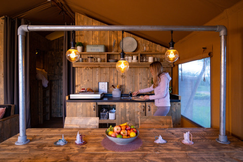 Una mujer prepara comida en la acogedora cocina de una tienda safari con muebles de madera e iluminación cálida.