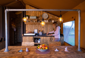 Une femme prépare un repas dans la cuisine chaleureuse d'une tente safari avec meubles en bois et lumière douce.