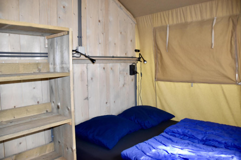 Interior de una tienda safari Comfort en Camping 't Veld, Países Bajos, con cama, estantería y ventana.