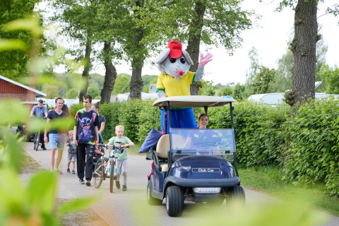En person klædt ud som musemaskot hilser fra en golfvogn foran familier på Camping 't Veld, Nederlandene.