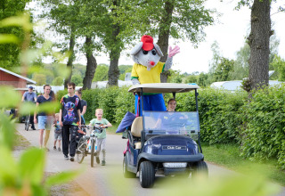 En person klædt ud som musemaskot hilser fra en golfvogn foran familier på Camping 't Veld, Nederlandene.