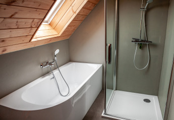Moderne badkamer met bad en douche onder schuin houten plafond in Heydehuis-lodge te Sandberghe.
