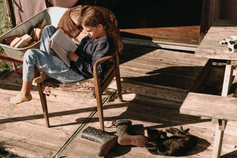 Niña leyendo en terraza de madera con gato y botas en Feather Down Hoeve De Pippert, Gelderland, Países Bajos.