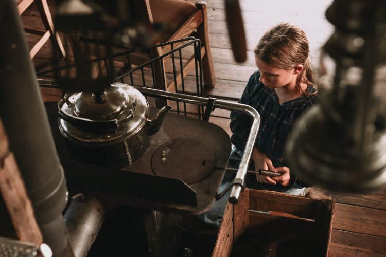 Child sits by an old wood stove with a kettle in a rustic cabin at Feather Down Hoeve De Pippert holiday park.