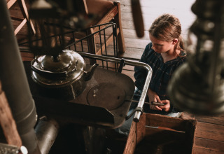 Child sits by an old wood stove with a kettle in a rustic cabin at Feather Down Hoeve De Pippert holiday park.