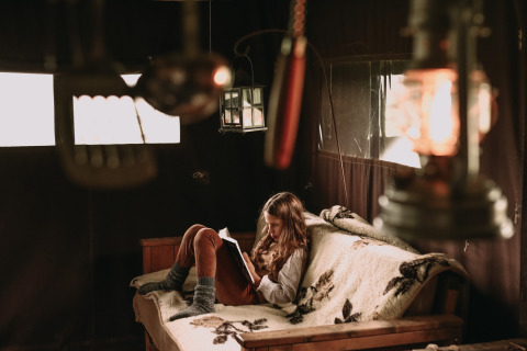 A girl reads a book on a blanket-covered sofa in a cozy, dimly lit cabin with warm ambient lanterns.