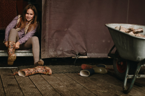 Une fille met des bottes en caoutchouc devant une tente, à côté d’une brouette pleine de bois.