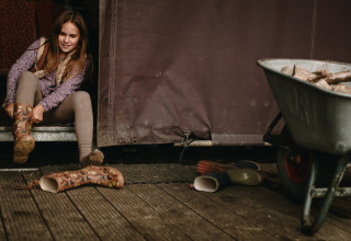 Une fille met des bottes en caoutchouc devant une tente, à côté d’une brouette pleine de bois.