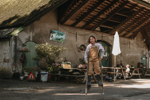 Enfant marchant sur des échasses devant une grange à Feather Down Hoeve De Pippert, parc de vacances aux Pays-Bas.