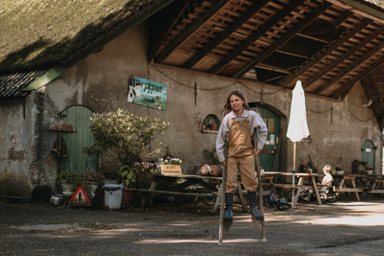 Child walking on stilts in front of rustic barn at Feather Down Hoeve De Pippert holiday park, Gelderland, Netherlands.