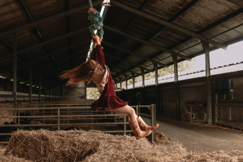 A girl in cowboy boots swings from a rope over hay in a barn at Feather Down Hoeve De Pippert, Gelderland.