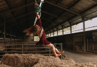A girl in cowboy boots swings from a rope over hay in a barn at Feather Down Hoeve De Pippert, Gelderland.
