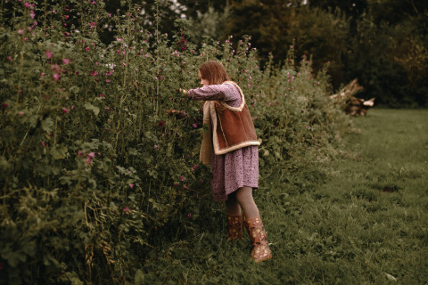 Een meisje in een paarse jurk en laarzen plukt bloemen in de tuin van Feather Down Hoeve De Pippert.