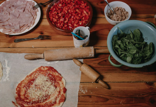 Forberedelse af pizza med skinker, tomater, spinat og andre ingredienser på et træbord i et feriehus.