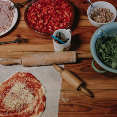 Preparando pizza con jamón, espinacas, tomates y más ingredientes en una mesa de madera en un parque vacacional.