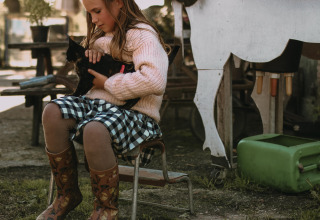 Girl in checkered dress and boots sits outdoors at Hoeve De Pippert, Gelderland, cuddling a black cat.