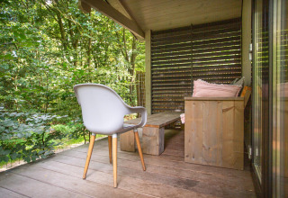 Veranda de madera en Boslodge De Thijmse Berg, Países Bajos, con silla moderna, banco y vista al bosque.