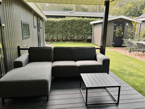 Zona de estar exterior con sofá gris y mesa en la terraza de una cabaña Bos Cottage Deluxe en los Países Bajos.