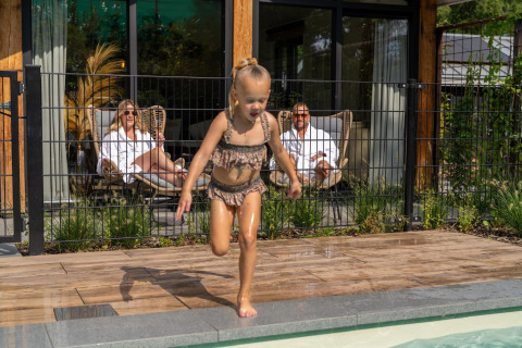 Kind speelt bij het zwembad terwijl twee volwassenen ontspannen aan het terras van Pool Lodge op De Thijmse Berg, Nederland.