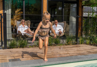 Kind speelt bij het zwembad terwijl twee volwassenen ontspannen aan het terras van Pool Lodge op De Thijmse Berg, Nederland.