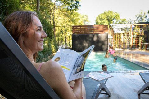 Mujer leyendo junto a la piscina en Pool Lodge, De Thijmse Berg, mientras niños juegan en el agua, Países Bajos.