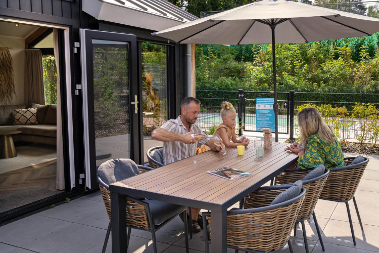 Familie entspannt sich beim Essen und Spielen unter Sonnenschirm in der Pool Lodge, De Thijmse Berg, Niederlande.