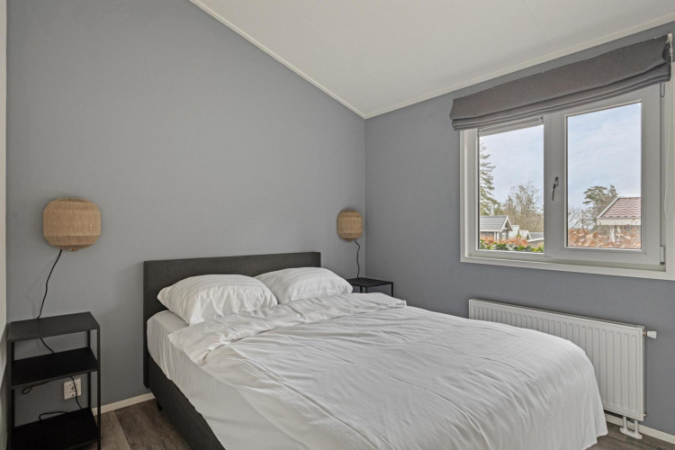 Dormitorio moderno con paredes grises, cama doble, ropa de cama blanca, mesillas y ventana grande en Wellness Lodge.