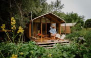 Een vrouw staat op het terras van een klein houten huisje, omringd door overvloedige natuur en planten.