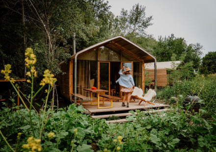 Een vrouw staat op het terras van een tiny house van hout, omringd door groene planten in de natuur.