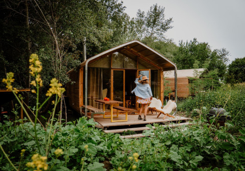 A woman stands on the deck of a tiny house in the woods, surrounded by lush greenery and wild plants.