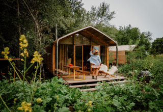Una mujer está en la terraza de una casa pequeña de madera, rodeada de vegetación y plantas silvestres.