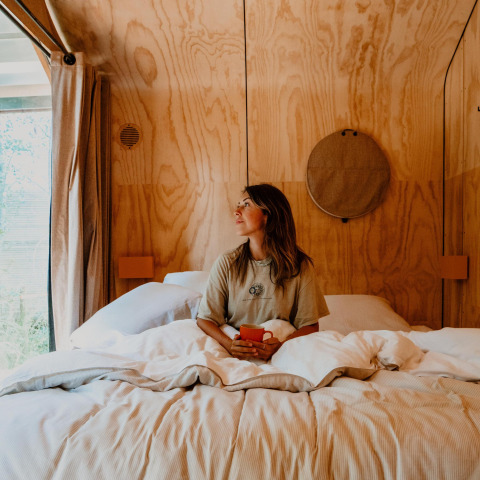 Vrouw zit op bed met een tas in een gezellige tiny house met houten muren en veel daglicht.