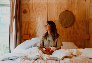 Mujer sentada en la cama con una taza en una tiny house acogedora con paredes de madera y luz natural.