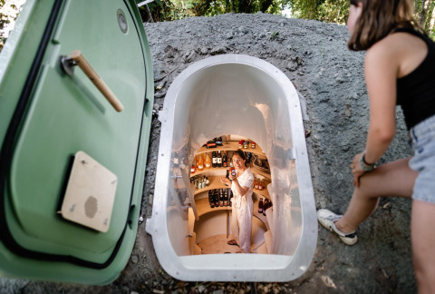 Vrouw kijkt in een ondergrondse kleine voorraadkelder bij een tiny house, andere vrouw lacht binnenin.