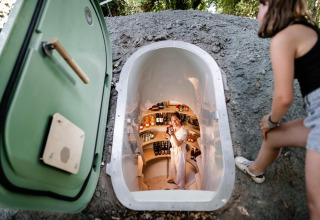 Vrouw kijkt in een ondergrondse kleine voorraadkelder bij een tiny house, andere vrouw lacht binnenin.
