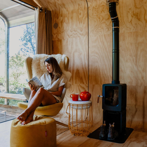 Vrouw leest in een comfortabele stoel in een tiny house met houtkachel, theepot en zonnig raam.