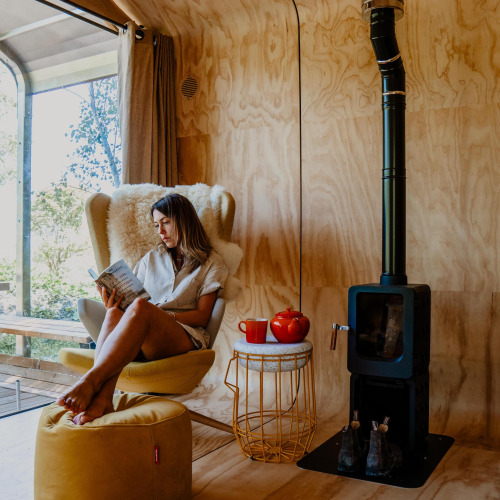Woman reads a book in a cozy armchair inside a tiny house with wood stove, tea set, and sunny view.