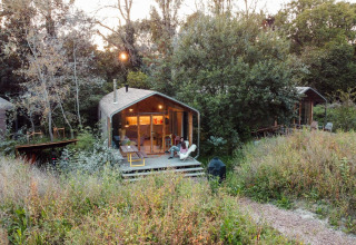 A tiny house in the woods, with a person relaxing on the porch surrounded by wild grass and trees.