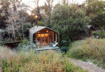 Tiny house immersa nella natura con una persona che si rilassa sul portico tra erba alta e alberi.