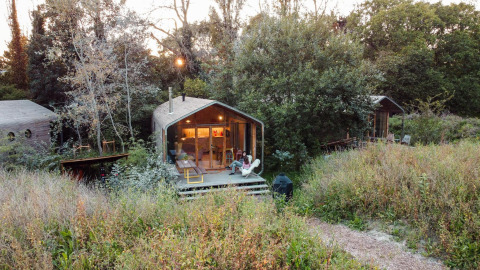 A tiny house in the woods, with a person relaxing on the porch surrounded by wild grass and trees.
