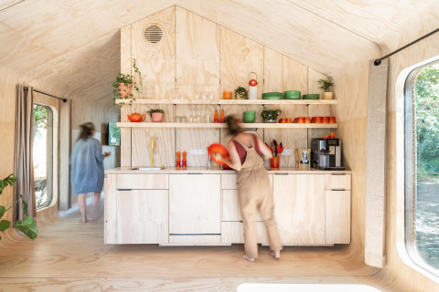 Twee personen in een lichte, moderne tiny house-keuken met houten afwerking en kleurrijk servies op open rekken.