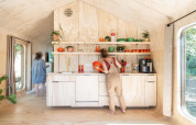 To personer i et lyst, moderne tiny house-køkken med trægulv og åbne hylder fyldt med farverige køkkenredskaber.