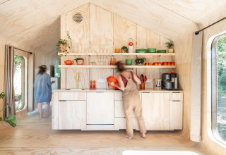Twee personen in een lichte, moderne tiny house-keuken met houten afwerking en kleurrijk servies op open rekken.
