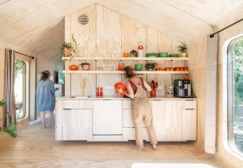 Dos personas en la cocina de una tiny house moderna y luminosa, con estanterías abiertas y vajilla colorida.