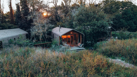 Pequeña casa rodeada de naturaleza y césped alto, con árboles y una terraza iluminada al atardecer.